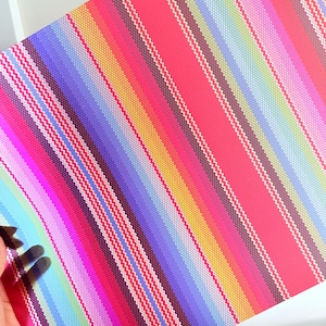 May include: A colorful, striped placemat with a woven texture. The design features diagonal stripes in shades of pink, red, purple, blue, green, yellow, and brown. The placemat is held by a hand.