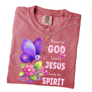May include: A red Comfort Colors t-shirt with a graphic of a purple butterfly, flowers, and the text "Blessed by GOD, Loved by JESUS, Led by the SPIRIT." A silver necklace with a pendant is also present.