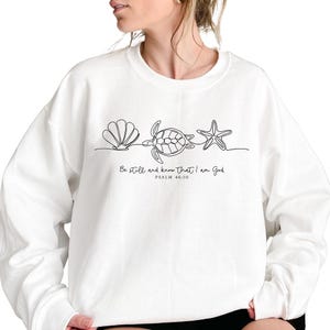 May include: White sweatshirt featuring a minimalist line drawing of a seashell, turtle, and starfish. Below the design is the text "Be still and know that I am God" and "Psalm 46:10" in black. The sweatshirt is a casual, comfortable style.
