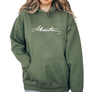 May include: An olive green hoodie with a front pocket and a drawstring hood. The hoodie features the white cursive text "A. Hamilton" across the chest. The garment is made of a soft material.
