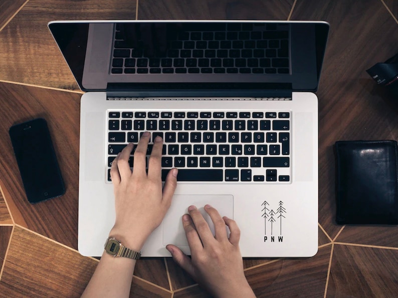 Pu&ograve; includere: Una persona sta digitando su un laptop argentato con una tastiera nera. Il laptop ha un adesivo nell'angolo in basso a destra che dice "PNW" con tre pini stilizzati. Il laptop &egrave; su una superficie di legno.