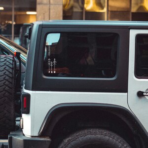 Pu&ograve; includere: Jeep Wrangler bianca con tetto nero e finiture nere. Il lunotto posteriore ha un adesivo con un disegno di tre alberi e le lettere "PNW".