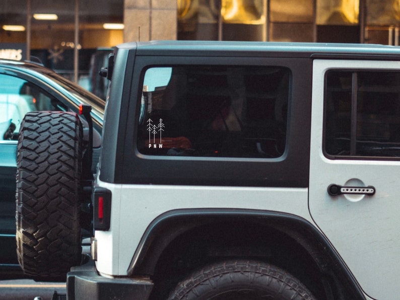 Pu&ograve; includere: Jeep Wrangler bianca con tetto nero e finiture nere. Il lunotto posteriore ha un adesivo con un disegno di tre alberi e le lettere "PNW".