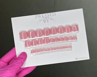 May include: A set of press-on nails with a pink base and white tips, arranged on a white card. The card has the text "PRESSED BY Emma" and a social media handle. The nails are in various sizes.