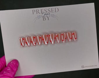 May include: A set of 10 pink press-on nails with a white line and red hearts design. The nails are displayed on a white card with the text "PRESSED BY" in black.