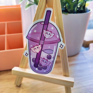 Puede incluir: Una pegatina morada y rosa de un vaso de boba tea con una pajita y tres gatos de dibujos animados rosa y blanco en su interior. La pegatina está en un pequeño caballete de madera.