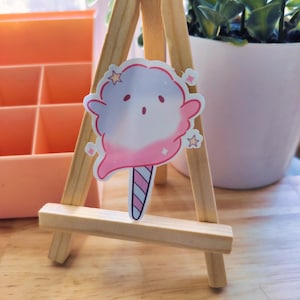 Puede incluir: Una pegatina de algodón de azúcar rosa y blanco con una linda carita de dibujos animados y un palo rayado rosa y blanco. La pegatina está en un pequeño caballete de madera.