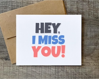 Könnte beinhalten: Eine weiße Grußkarte mit dem Text "HEY, I MISS YOU!" in Grau, Blau und Koralle. Die Karte liegt auf einer Holzoberfläche, mit einem braunen Umschlag im Hintergrund. Eine einfache Karte, um das Vermissen auszudrücken.