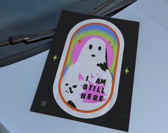 May include: Black rectangular sticker featuring a white ghost illustration with the text "I AM STILL HERE" in pink and white. The ghost is set against a rainbow gradient background within a white and orange oval. Yellow star accents are on the corners.