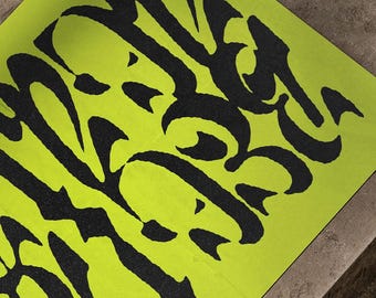 May include: A neon green rectangular paper with black abstract calligraphy. The calligraphy is stylized and covers most of the paper's surface. The paper is angled on a brown surface.