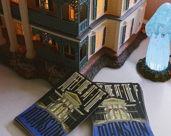 May include: A detailed, illuminated model of a mansion with a dark roof, intricate balconies, and glowing windows. Several cards with the text "MEET ME AT THE MANSION" are stacked in front of the mansion.