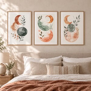 May include: A set of three framed wall art prints featuring watercolour illustrations of crescent moons, circular shapes, and botanical leaf sprigs in shades of terracotta orange, sage green, and dark charcoal grey on a light cream background.