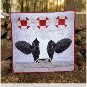 May include: A white and red patchwork quilt with a black and white cow face peeking out from the center. The quilt is hanging on a clothesline in a forest setting. The quilt is a pattern collaboration between Red Spool Designs and createdanew designs.