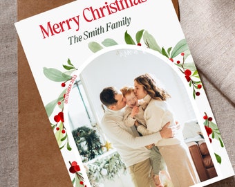 May include: A Christmas card with a white background and a green and red holly border. The card features a photo of a family of three, a man, a woman, and a child, standing in front of a Christmas tree. The text "Merry Christmas The Smith Family" is printed in red at the top of the card.