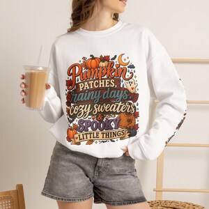 May include: A white long-sleeve sweatshirt featuring a colourful graphic print with the text Pumpkin Patches, rainy days, cozy sweaters & SPOOKY LITTLE THINGS, surrounded by illustrations of pumpkins, autumn leaves, a ghost, and a sweater.