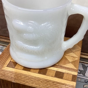 May include: A white, vintage-style mug with a dragon design on the side. The mug has a sturdy handle and sits on a wooden coaster with a geometric pattern. The mug is made of opaque glass.