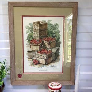 May include: Framed watercolor print featuring wooden baskets overflowing with ripe red strawberries. The artwork includes a stack of empty baskets and lush green foliage. The print is set within a light brown wooden frame.