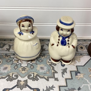 May include: A pair of vintage ceramic salt and pepper shakers. One shaker is a woman wearing a blue-trimmed hat and a white dress. The other shaker is a man wearing a blue-trimmed hat and a white outfit with a blue scarf. Both have brown hair and painted facial features.