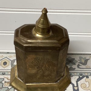 May include: A vintage brass box with an octagonal body and a conical lid. The box features etched designs, including a bird or insect motif. The box sits on a matching octagonal base, showcasing an antique aesthetic.