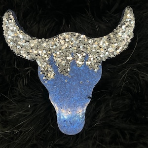 Puede incluir: Un adorno de cráneo de toro azul y plateado con purpurina. El cráneo está hecho de madera y tiene un acabado desgastado. Los cuernos están cubiertos de purpurina plateada y el cráneo está cubierto de purpurina azul.