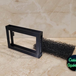 May include: A black plastic filter holder with a black foam filter. The filter holder is rectangular with a rectangular opening. The foam filter is rectangular and has a textured surface. The text "Custom Aquatics" is printed on a black circle with green text.