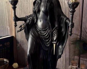 Könnte beinhalten: Schwarze Statue einer bekleideten Figur, die Fackeln mit goldenen Flammen hält. Die Figur trägt eine Halbmondkrone und hält einen Dolch und Schlüssel. Die Statue steht auf einer schwarzen Kiste mit Schädeldetail.