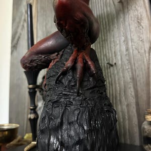 May include: A dark, detailed sculpture of a mythical creature with red and brown tones. The figure is perched on a textured black base, with a focus on the creature's clawed feet and body. The sculpture has a gothic or fantasy theme.