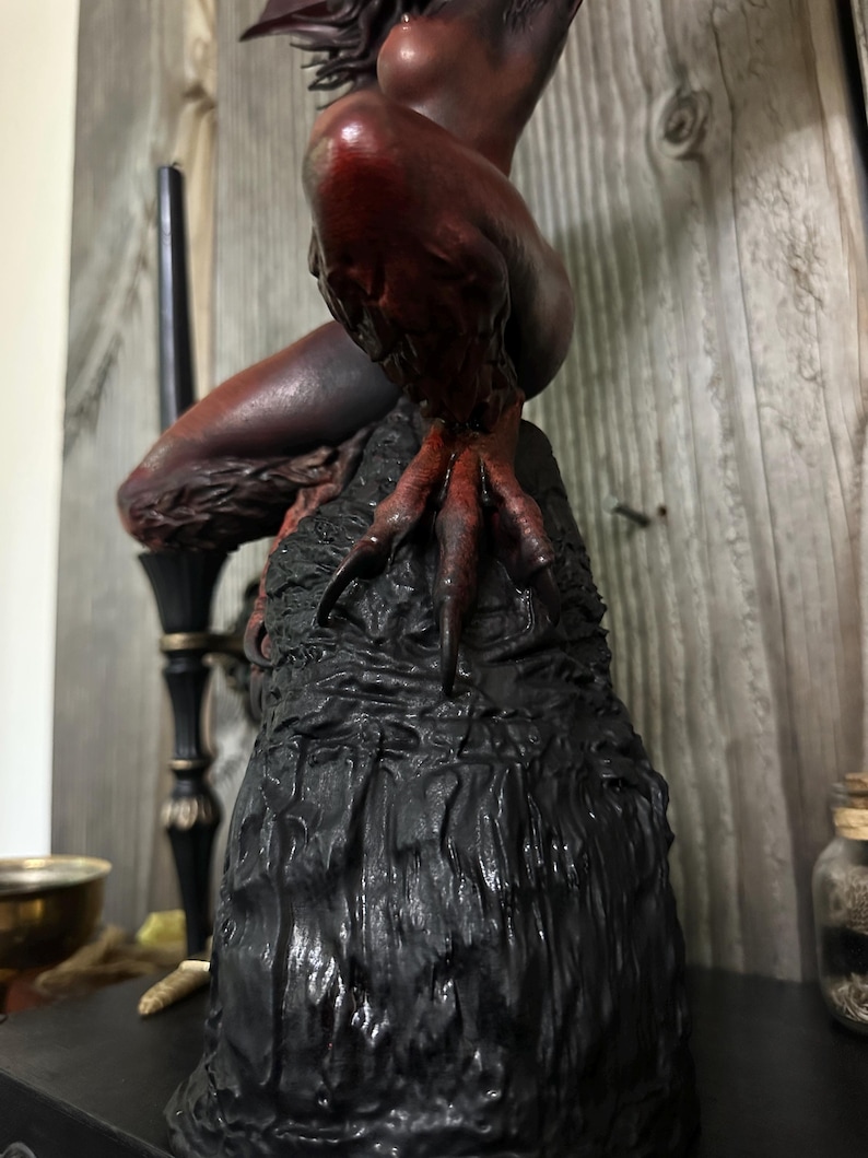 May include: A dark, detailed sculpture of a mythical creature with red and brown tones. The figure is perched on a textured black base, with a focus on the creature's clawed feet and body. The sculpture has a gothic or fantasy theme.