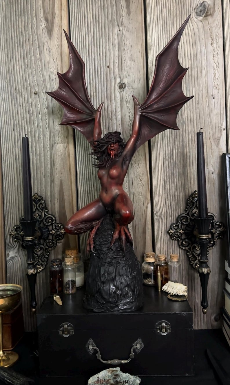 May include: A dark red and brown sculpture of a demonic figure with large wings, posed on a black base. The figure has a long tongue and clawed feet. The sculpture is displayed on a black box with decorative candle holders on either side.