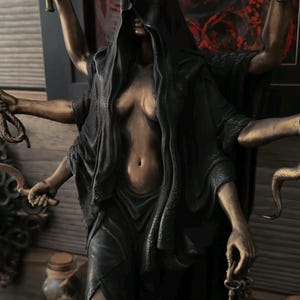 May include: A multi-armed bronze-toned statue of a figure wearing a black hooded cloak and a crown with a crescent moon. The figure holds two torches, a rope, a curved blade, and a set of keys in its six hands. The statue features a dark finish.