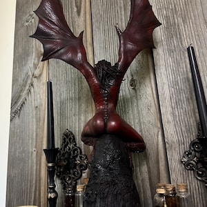 May include: A red and black sculpture of a winged figure in a kneeling position. The figure has large, outstretched wings and is perched atop a textured, black base. The background includes wooden paneling and decorative elements.