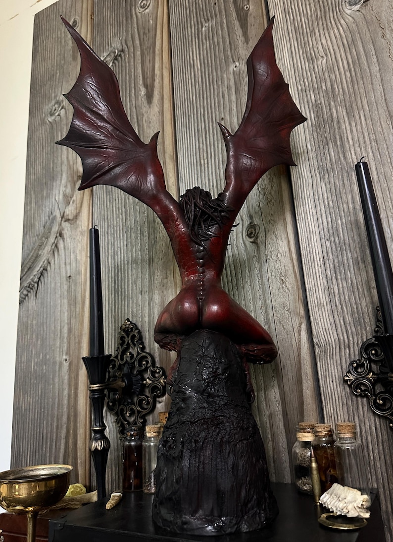 May include: A red and black sculpture of a winged figure in a kneeling position. The figure has large, outstretched wings and is perched atop a textured, black base. The background includes wooden paneling and decorative elements.