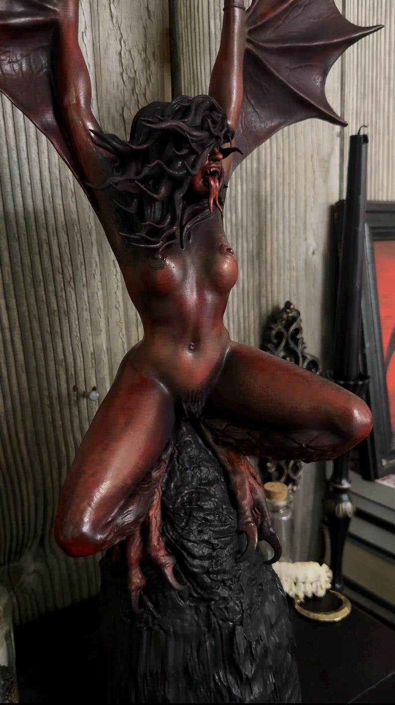 May include: A red and black sculpture of a demonic figure with bat wings, posed in a kneeling position. The figure has a snarling expression, long tongue, and is perched on a textured base. The sculpture is detailed with a dark, gothic aesthetic.