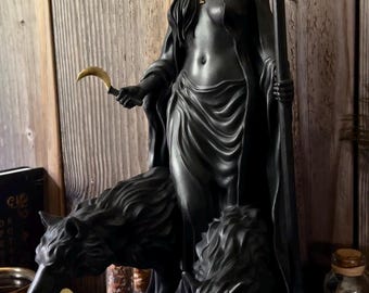Könnte beinhalten: Schwarze Statue einer bekleideten Figur mit goldener Krone und Fackel, die eine Sichel hält, flankiert von zwei Wolfsfiguren. Die Statue steht auf einer schwarzen Truhe vor einem hölzernen Hintergrund.