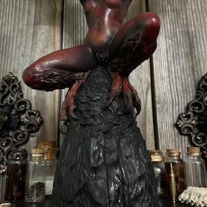 May include: A dark red and black sculpture of a demonic figure with bat-like wings, perched atop a textured, black base. The figure has sharp claws and a menacing expression. The sculpture is detailed and appears to be handmade.