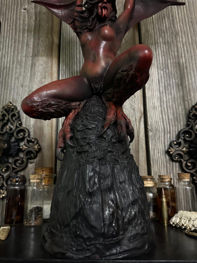May include: A dark red and black sculpture of a demonic figure with bat-like wings, perched atop a textured, black base. The figure has sharp claws and a menacing expression. The sculpture is detailed and appears to be handmade.