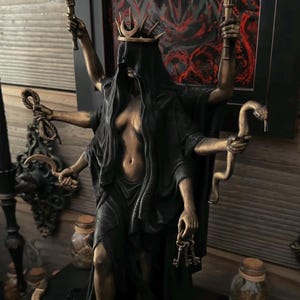 May include: A bronze-toned multi-armed statue of a figure wearing a black veil and a crown with a crescent moon. The figure holds two flaming torches, a rope, a curved blade, a snake, and a set of keys. The statue features a dark, flowing robe.