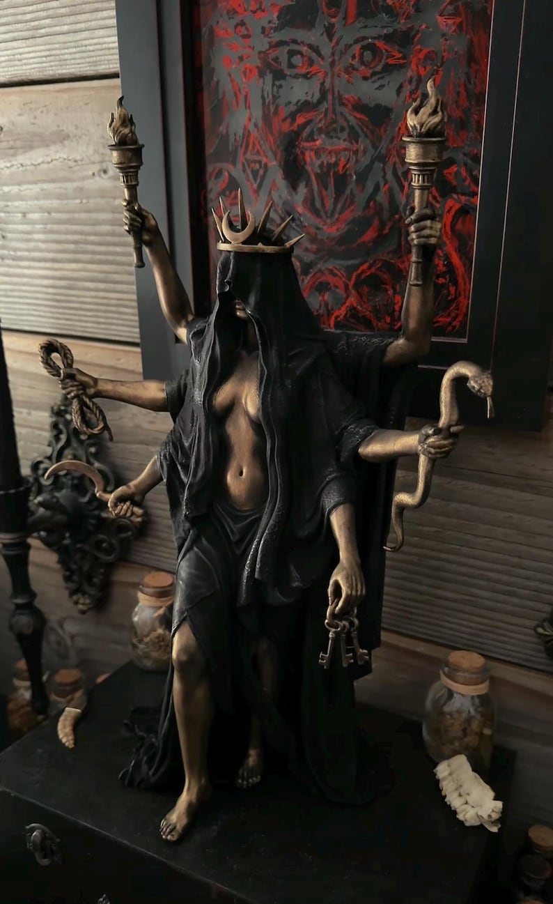 May include: A bronze-toned multi-armed statue of a figure wearing a black veil and a crown with a crescent moon. The figure holds two flaming torches, a rope, a curved blade, a snake, and a set of keys. The statue features a dark, flowing robe.