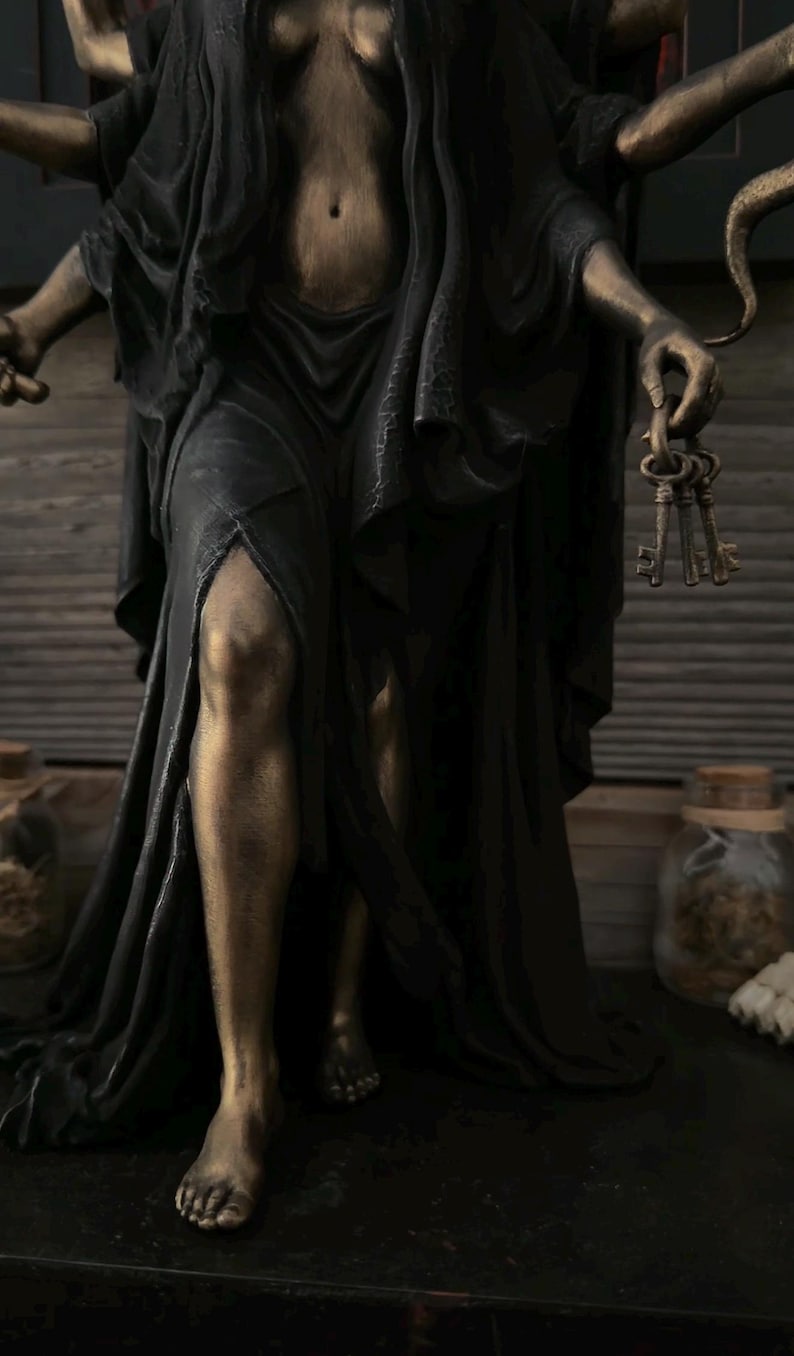 May include: A multi-armed statue with a bronze-toned finish, wearing a flowing black garment with a high leg slit. The figure has multiple arms, one of which holds a set of three metal keys. The statue is depicted in a standing position.