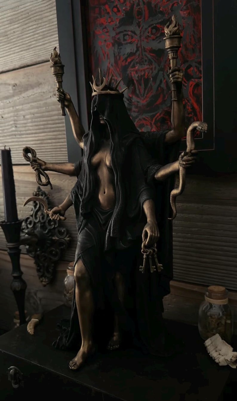 May include: A bronze-toned multi-armed statue of a figure wearing a black hooded cloak and a crescent moon crown. The figure holds two torches, two snakes, a curved blade, and a set of keys in its six hands, with a dark, draped garment.