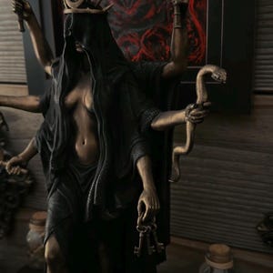 May include: A bronze-toned multi-armed statue wearing a black hooded cloak and a crescent moon crown. The figure holds two flaming torches, a coiled snake, and a set of keys. The statue has a dark, weathered finish and stands in a dynamic pose.