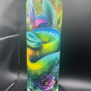 May include: A cylindrical tumbler featuring a vibrant illustration of a colorful snake coiled amidst lush tropical foliage. The design incorporates shades of blue, green, yellow, and purple. The tumbler sits on a white base.