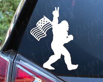 May include: A white vinyl sticker of a Bigfoot holding an American flag with a peace sign. The sticker is on the rear window of a silver car.