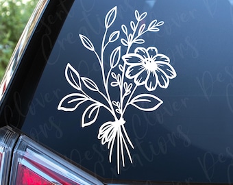 May include: A white vinyl decal featuring a line art illustration of a wildflower bouquet tied with a bow, applied to the rear window of a vehicle. The design consists of a central daisy-like flower surrounded by various leaves and stems.