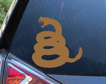 May include: A tan-colored vinyl decal of a coiled rattlesnake with its mouth open, displayed on the rear window of a vehicle. The design features the iconic silhouette of a snake in a defensive posture with a visible rattle at the tail end.
