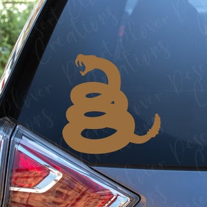 Op de afbeelding: Een beige vinyl sticker van een opgerolde ratelslang met open bek, aangebracht op de achterruit van een voertuig. Het ontwerp toont het iconische silhouet van een slang in een defensieve houding met een zichtbare ratel aan het uiteinde.