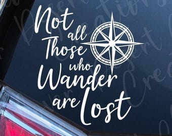 May include: A white vinyl decal applied to a car window featuring a compass rose graphic and the text "Not all those who wander are lost" in a stylized script font. The design is composed of clean white lines against the dark glass surface.