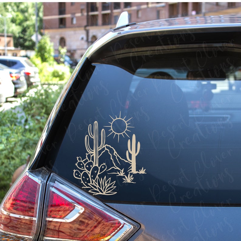 May include: A gold outline of a desert scene with a sun, mountains, cacti, and prickly pear plants. The design is perfect for a car window decal or sticker.