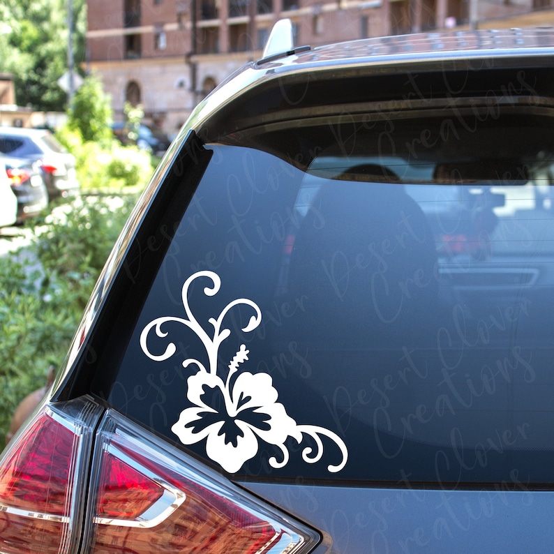 May include: White vinyl decal of a hibiscus flower with leaves and vines. The decal is applied to the back window of a dark blue car.