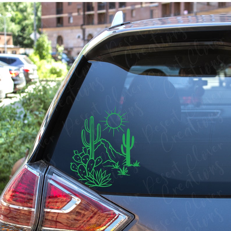 May include: Green vinyl decal of a desert scene with cacti, a mountain, and a sun. The decal is on the back window of a silver car.